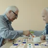 Dwóch starszych mężczyzn gra w grę dla seniorów mindalo przy stoliku z filiżankami kawy. Jeden z nich odwraca kartę z pomarańczowym symbolem, angażując się w trening pamięci.