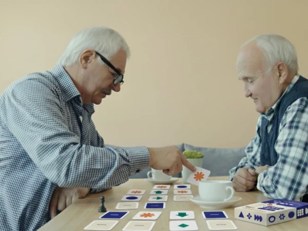 Dwóch starszych mężczyzn gra w grę dla seniorów mindalo przy stoliku z filiżankami kawy. Jeden z nich odwraca kartę z pomarańczowym symbolem, angażując się w trening pamięci.